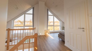 A spacious room with a high ceiling, large windows, and a wooden staircase. - Newly built vacation home with many highlights (sauna, fireplace and first lake location) (Nordhorn)