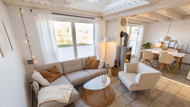 A modern living room with a sofa, armchair, and coffee table, a dining area with a table and chairs, and a fireplace. - Newly built vacation home with many highlights (sauna, fireplace and first lake location) (Nordhorn)