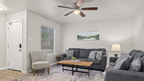 Un salon avec un canapé gris, une table basse en bois, un fauteuil gris, un ventilateur de plafond et un tableau accroché au mur.