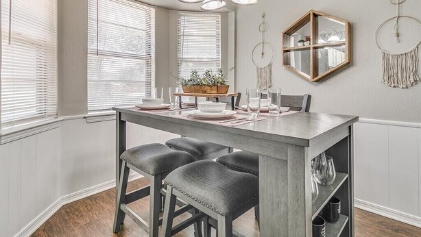 Un coin repas avec une table en bois, quatre chaises et un lustre. On y trouve des fenêtres avec des stores, un miroir et des éléments de décoration murale.