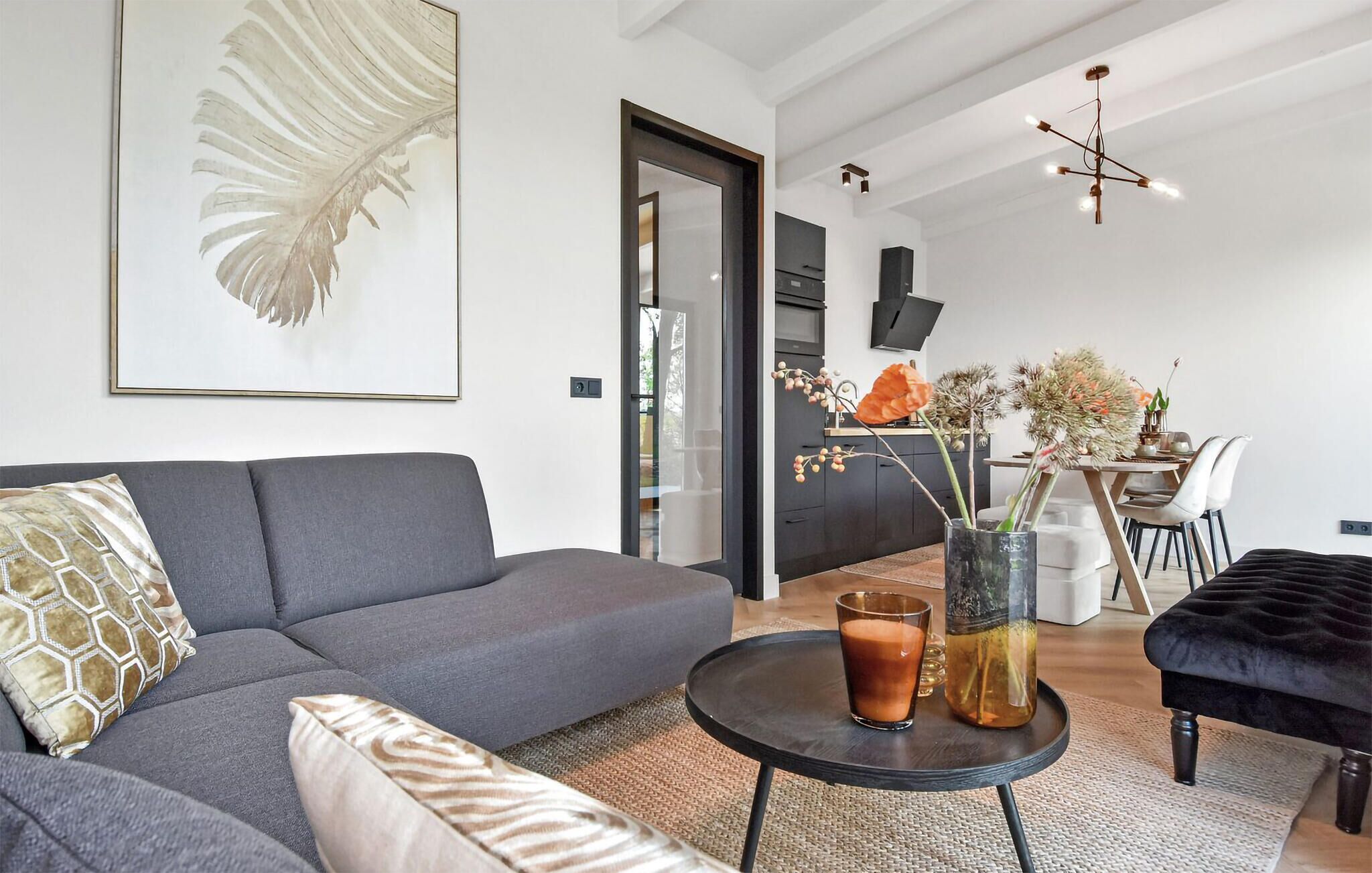 A modern living room with a grey sofa, a round coffee table, and a black ottoman. There is a dining area with a table and chairs visible in the background.