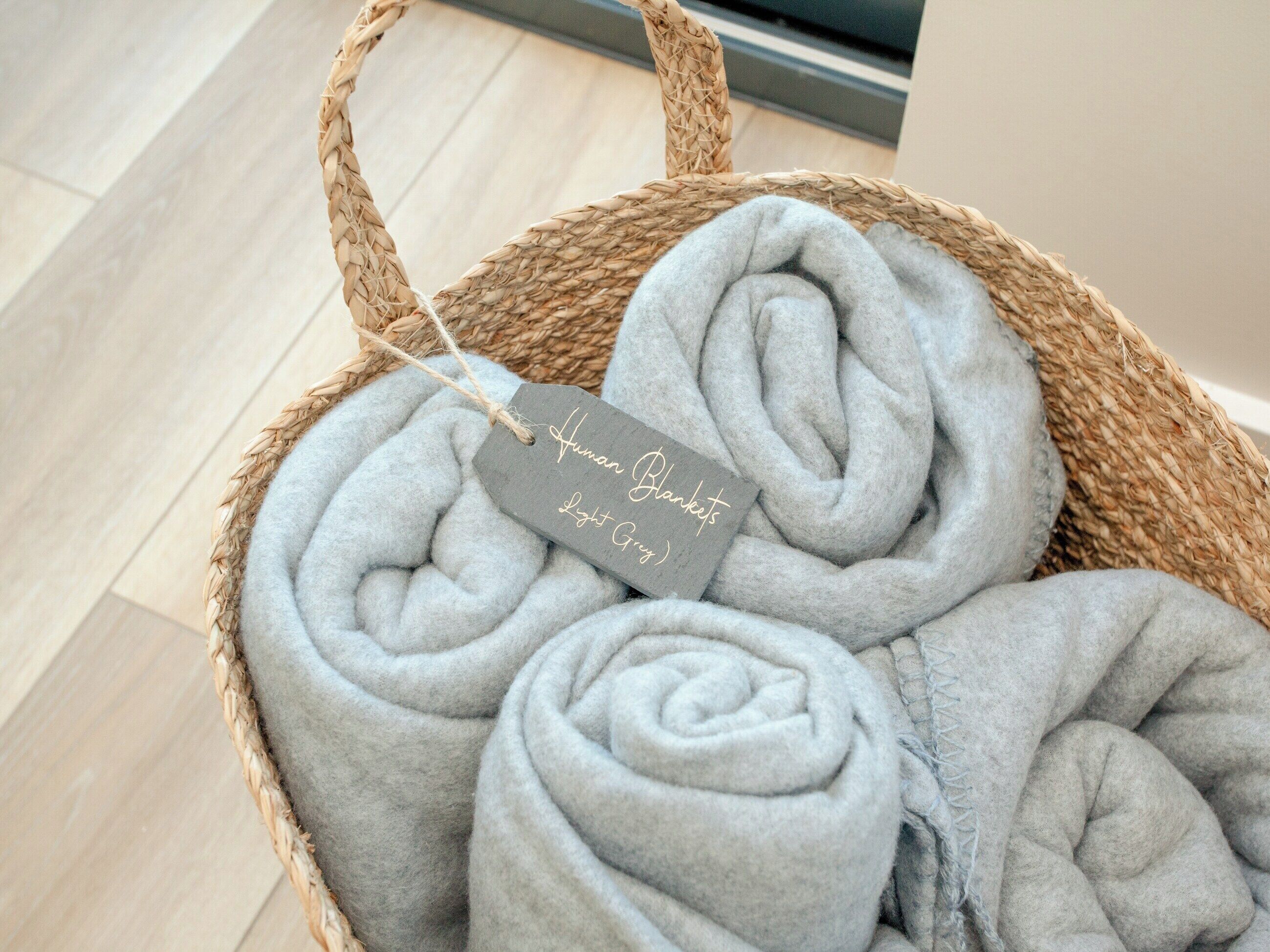 Una cesta tejida con mantas grises enrolladas, con una etiqueta que dice 'Human Blankets Light Grey'.