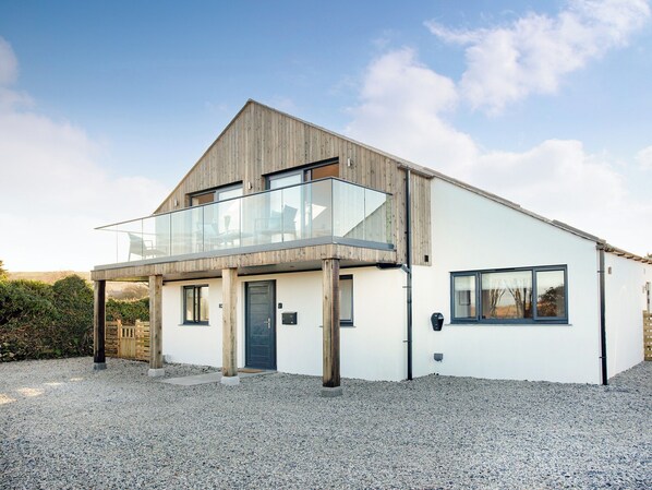 Et moderne hus i to etager med en træfacade, en balkon med gelændere i glas og en grusbelagt indkørsel.