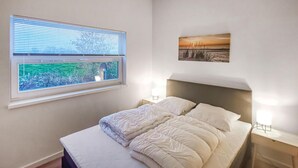Un dormitorio con cama, ventana con persianas, mesita de noche con lámpara y un cuadro en la pared.