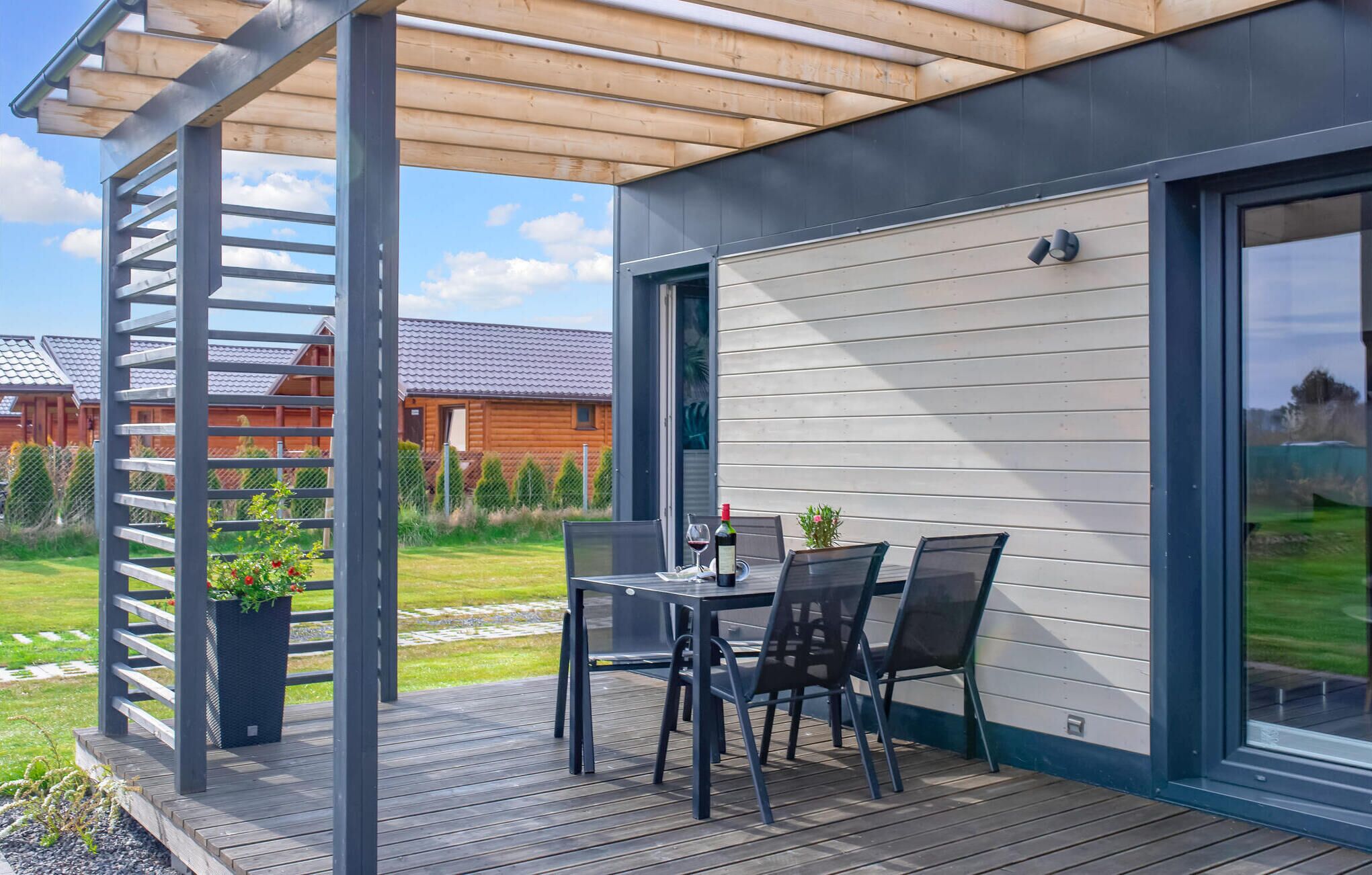 A covered outdoor patio with a table and chairs, a pergola, and a view of a wooden house in the background.