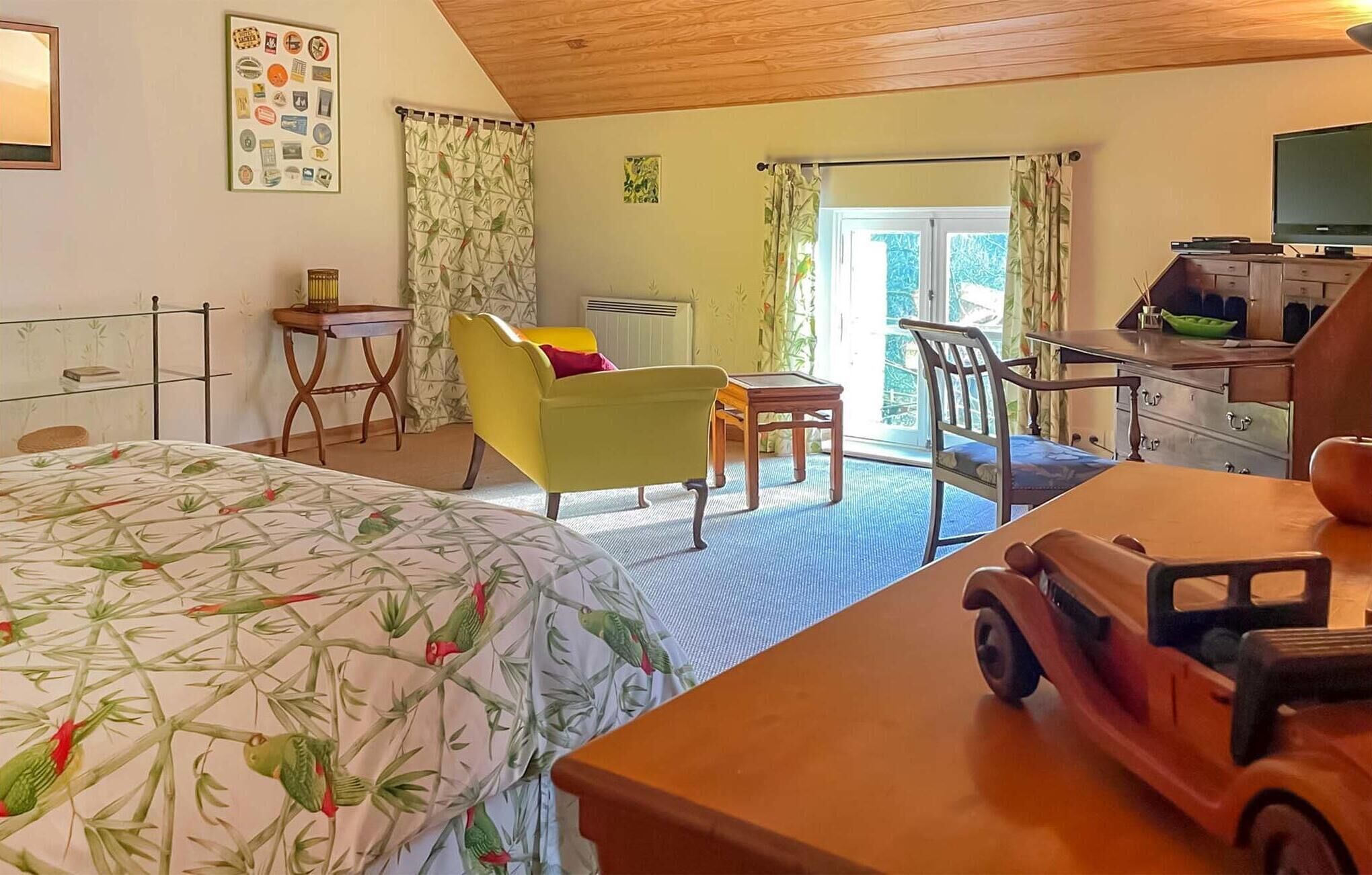 A room with a bed, a yellow armchair, a wooden table, a television, and a small toy car.