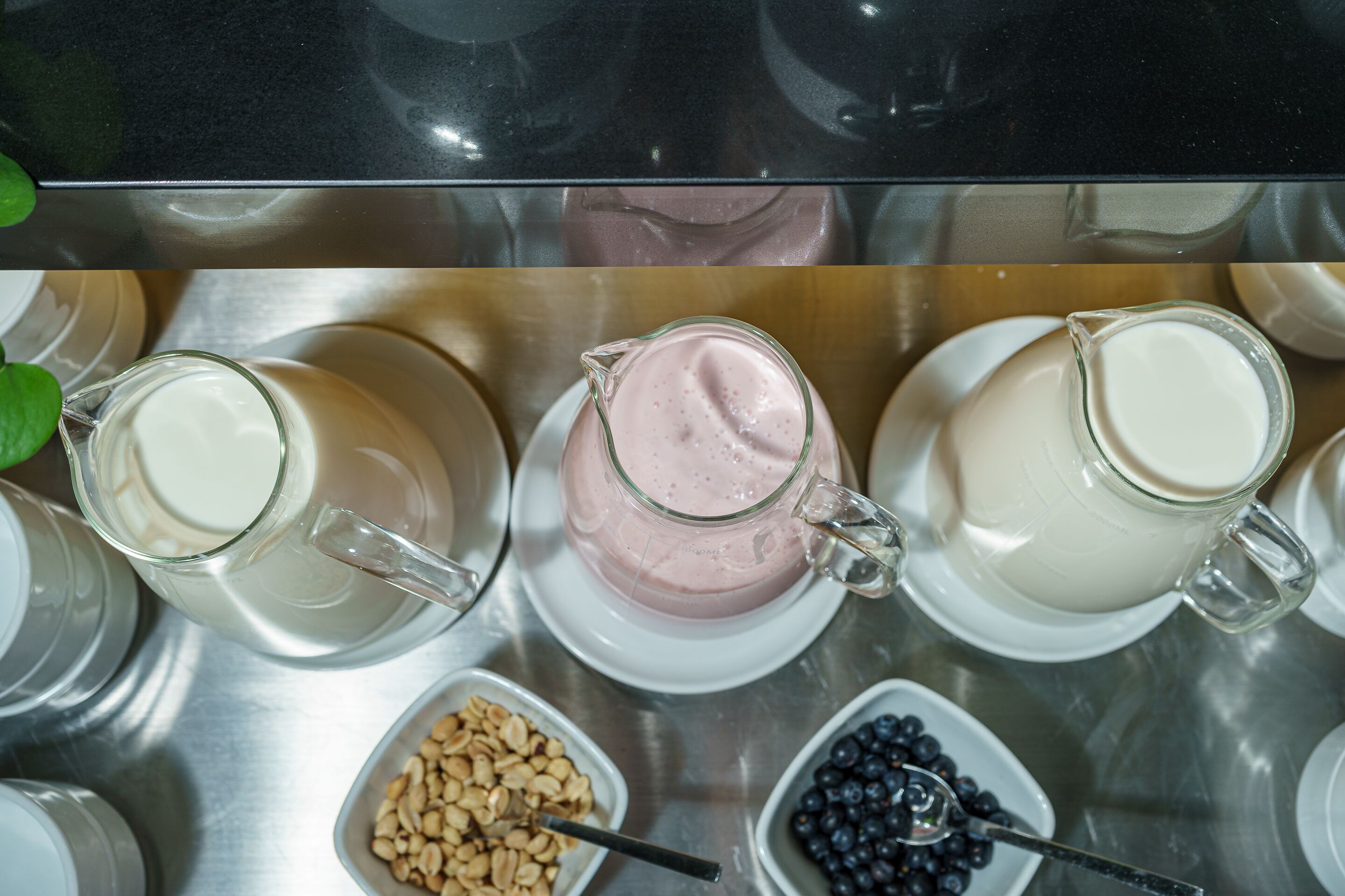 A stainless steel table with three glass pitchers of milk and two bowls of granola and blueberries.