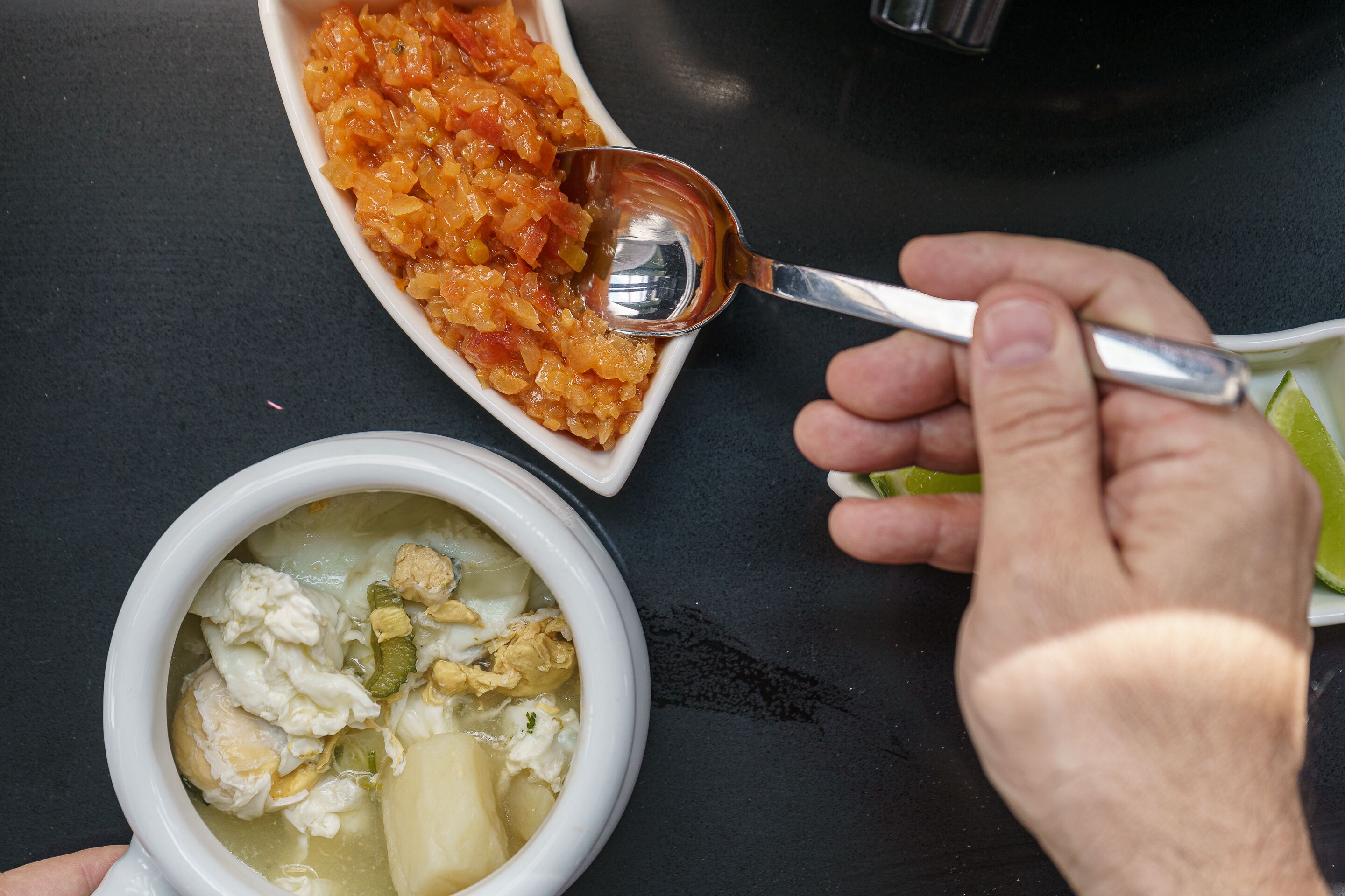 Un tazón de sopa con una cuchara, un pequeño recipiente de salsa y una cuña de lima sobre una mesa.