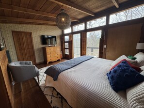 A bedroom with a large bed, wooden ceiling, a television, and a view through the windows.