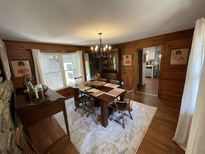 A dining room with a wooden table, chairs, and a chandelier. There are framed pictures on the walls and a window with curtains.