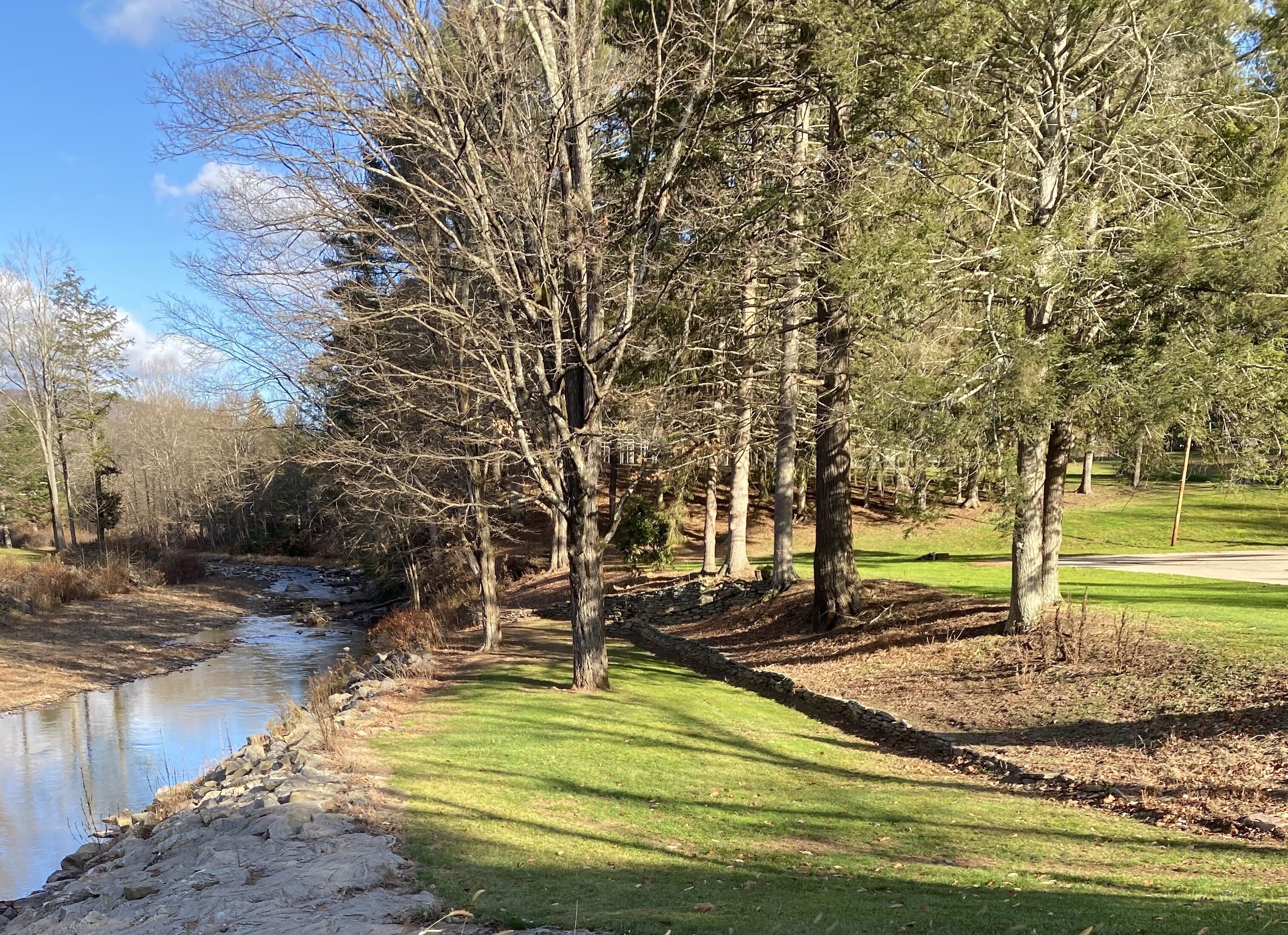 Een rustig pad langs de rivier, met een stenen muur en grasvelden, omgeven door bomen.