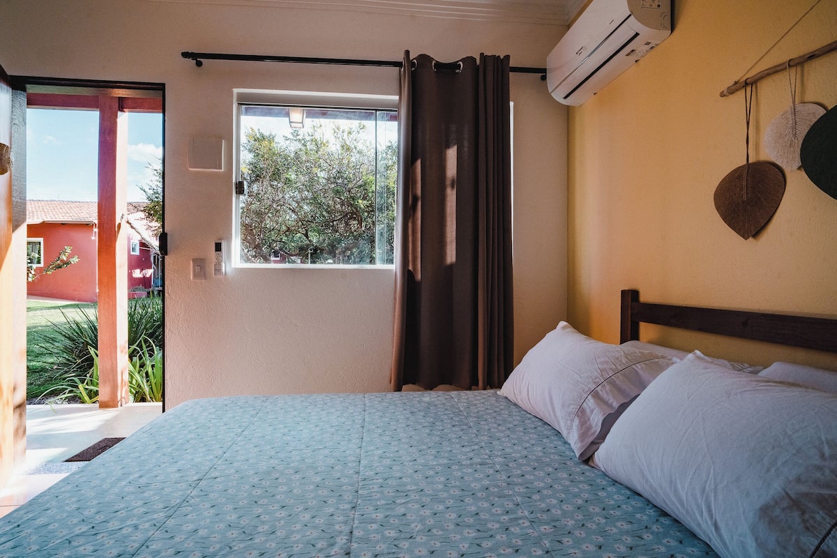 Un dormitorio con cama, ventana, aire acondicionado y vista a un edificio rojo.