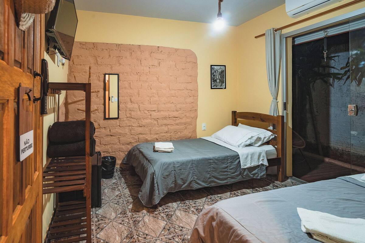 Una habitación con dos camas, una silla de madera, una mesita, un muro de ladrillo y una ventana con cortinas.