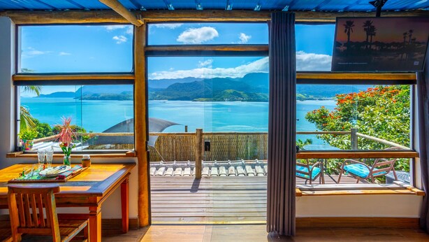 A room with large windows offering a view of the sea and mountains. There is a wooden table with chairs and a small balcony with a chair.