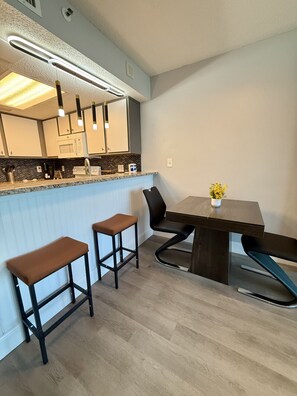 A modern kitchen with a bar area, two stools, a dining table, and a vase with flowers.
