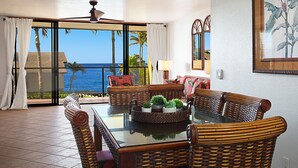 A dining area with a glass-top table surrounded by wicker chairs, a view of the ocean, and a ceiling fan. - Recently upgraded single level ocean view condo (Koloa)