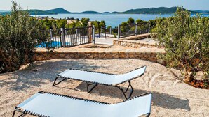 Un espacio al aire libre iluminado por el sol con dos sillones, un sendero de piedra y vista a un lago y colinas al fondo.