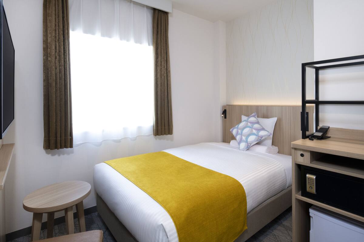 Hotellihuone, jossa on sänky, yöpöytä, lamppu ja ikkuna, jossa on verhot.