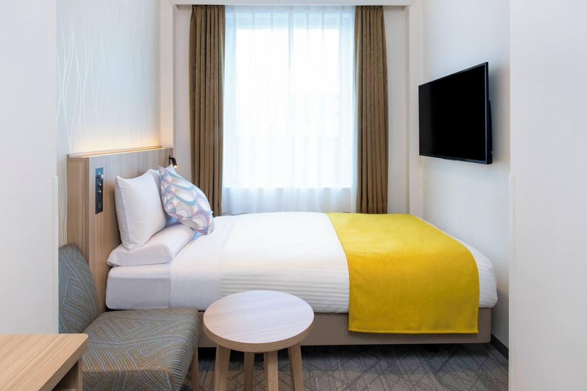 Hotellihuone, jossa on sänky, yöpöytä, pieni pyöreä pöytä, seinälle asennettu televisio ja ikkuna, jossa on verhot.