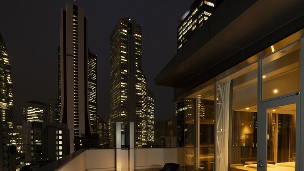 Un edificio alto con finestre illuminate di notte, un balcone con pavimento in legno e una stanza vetrata con un letto e angolo salotto.