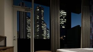 Camera d'albergo con un letto, un piccolo tavolo rotondo, una panca e vista sugli edifici della città di notte.