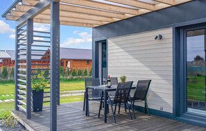 Une terrasse extérieure couverte, avec une table et des chaises, une pergola et une vue sur une maison en bois à l’arrière-plan.