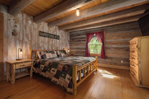 A log cabin bedroom with a bed, wooden nightstands, a dresser, and a window with red curtains. - "Timber Lodge", Rustic 4BR Log Cabin + Guesthouse (Bluff City)