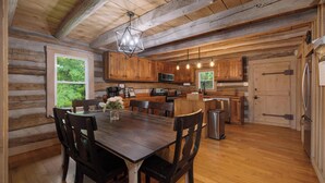 A rustic cabin interior with a wooden dining table, chairs, a kitchen area with wooden cabinets, a hanging light fixture, and a view of greenery outside. - "Timber Lodge", Rustic 4BR Log Cabin + Guesthouse (Bluff City)