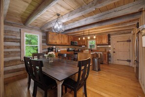 Ein rustikales Blockhaus-Interieur mit einem Holzespeisesaal, Stühlen, einer Küchenzeile mit Holzausstattung, einer Pendelleuchte und Blick ins Grüne.