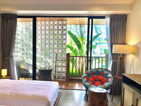 A hotel room with a bed, a chair, a table, a lamp, and a view of greenery outside.