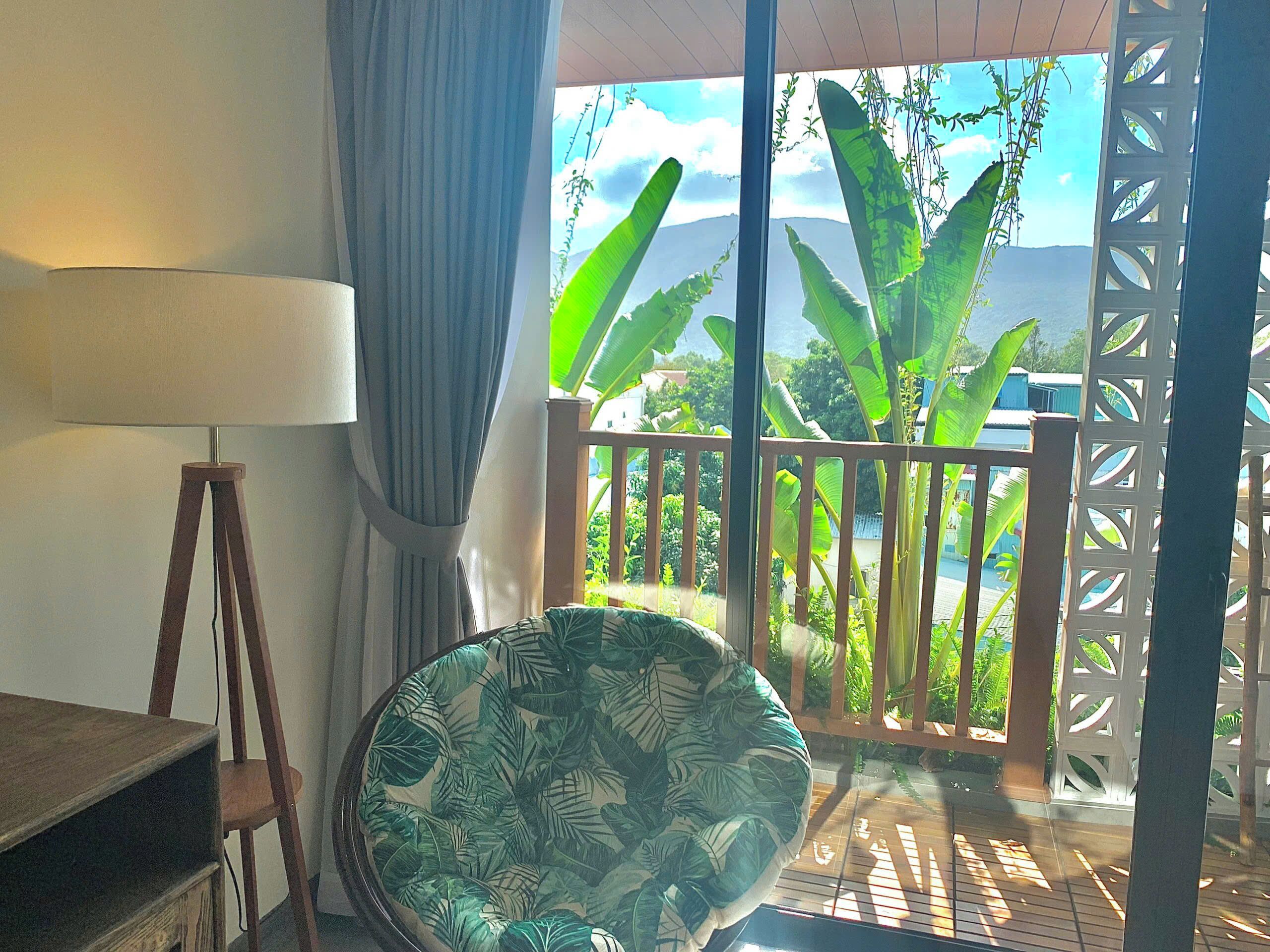 A room with a wooden floor, a round chair with a leaf pattern, a lamp, and a view of greenery through a balcony.