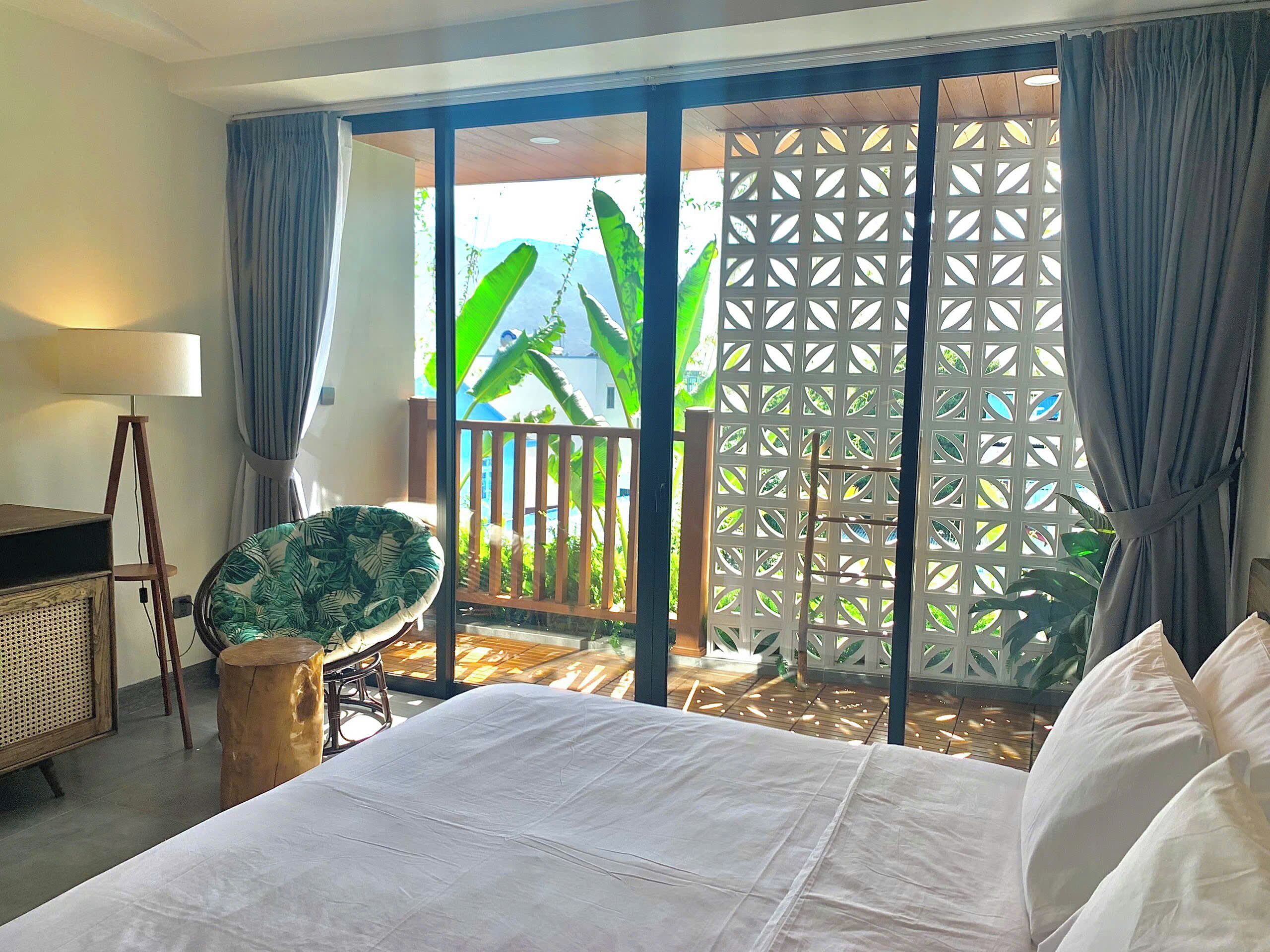 A hotel room with a bed, a chair, a lamp, and a view of a balcony with plants.