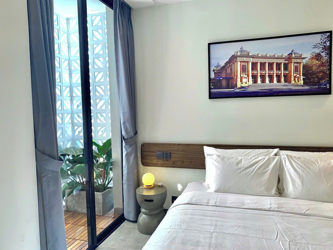 A hotel room with a bed, a nightstand with a lamp, a painting on the wall, and a view of a building through the window.