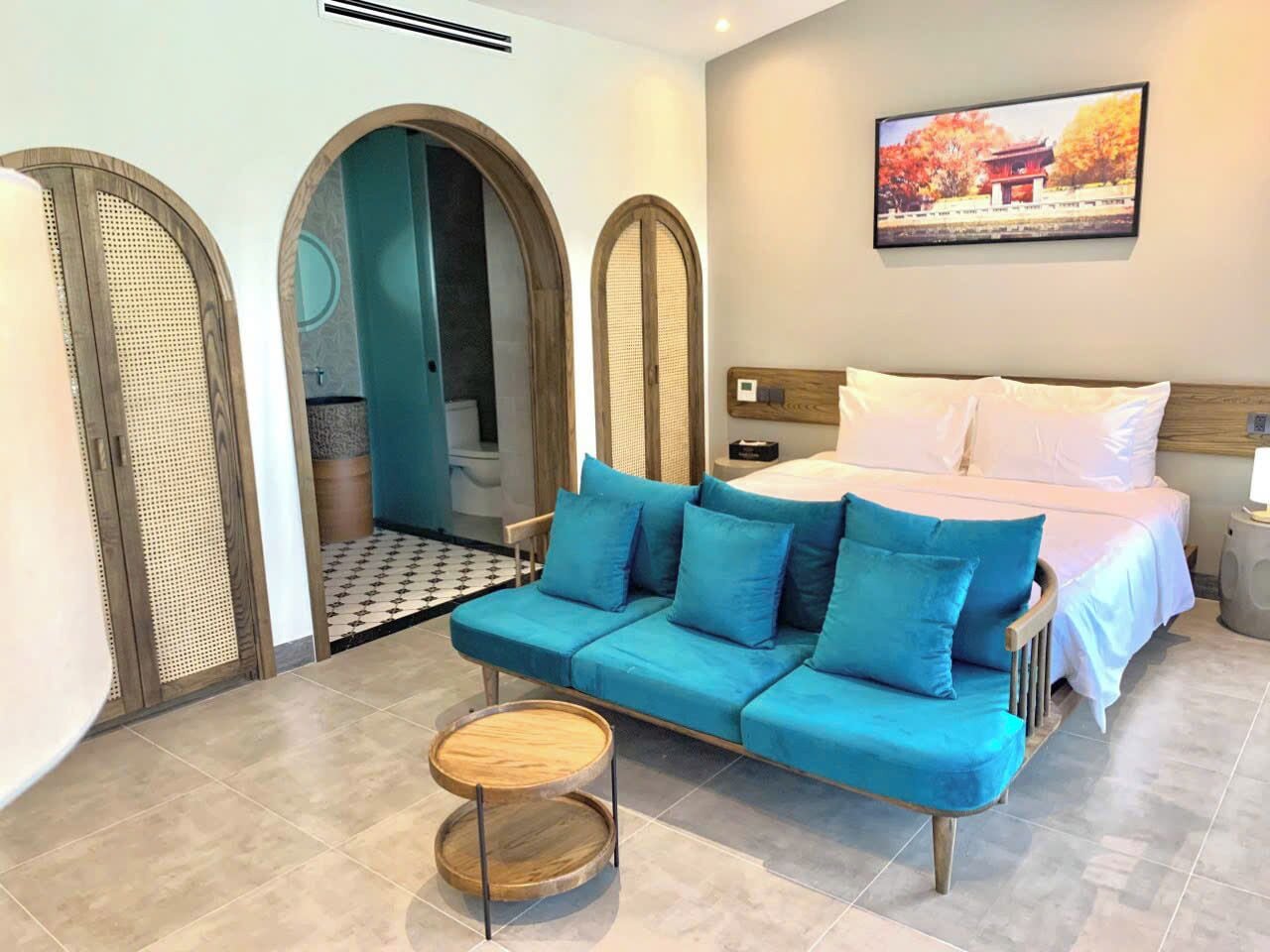 Quarto de hotel moderno com uma cama grande, um sofá azul, uma mesa redonda pequena e vista para um edifício tradicional pela janela.