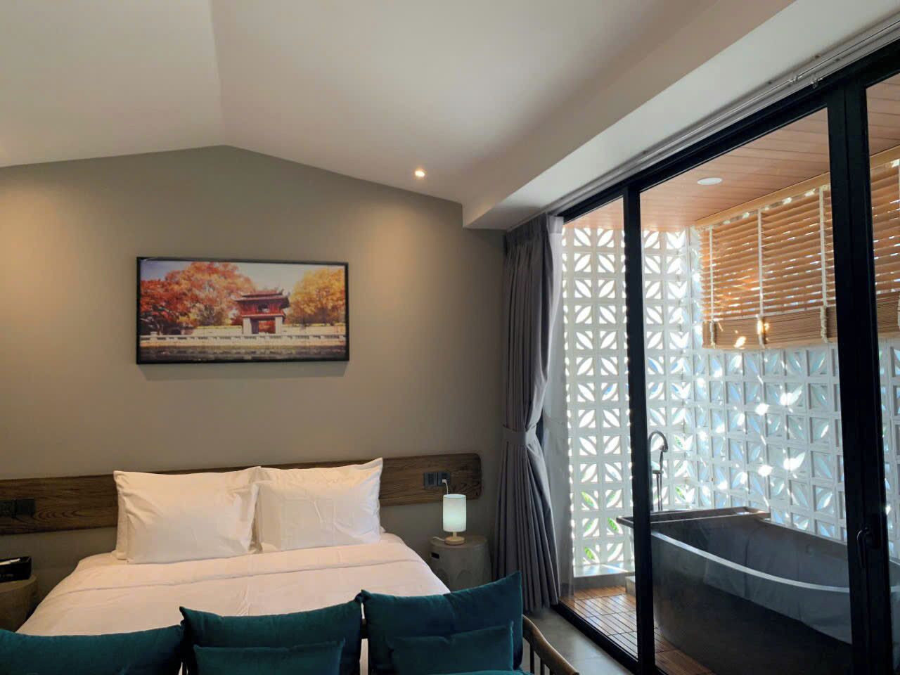 A hotel room with a bed, a nightstand, a wall-mounted lamp, a window with a view of a balcony, and a painting on the wall.