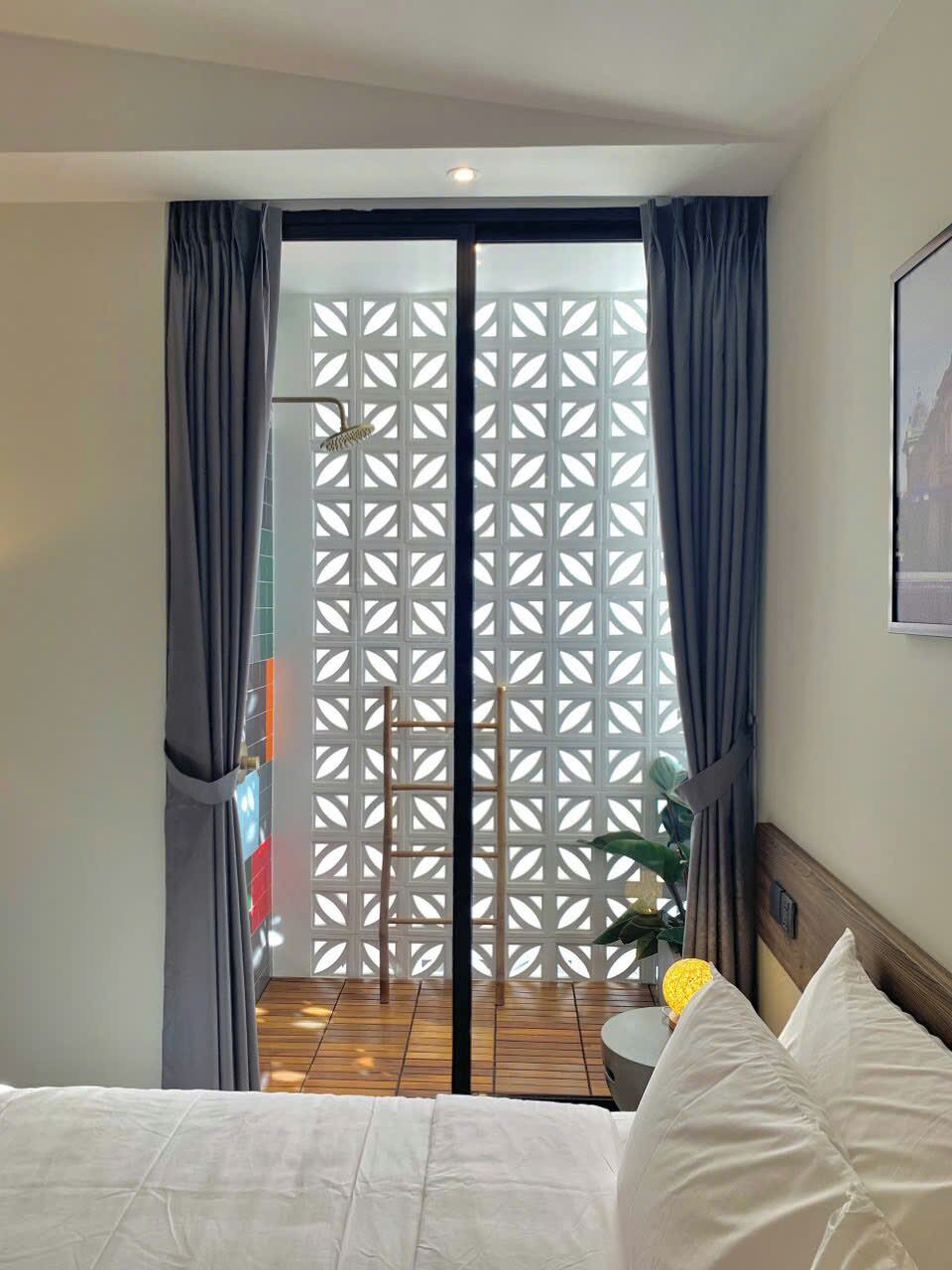 A hotel room with a bed, a small round table with a lamp, a wooden floor, and a view through a window with a lattice screen.