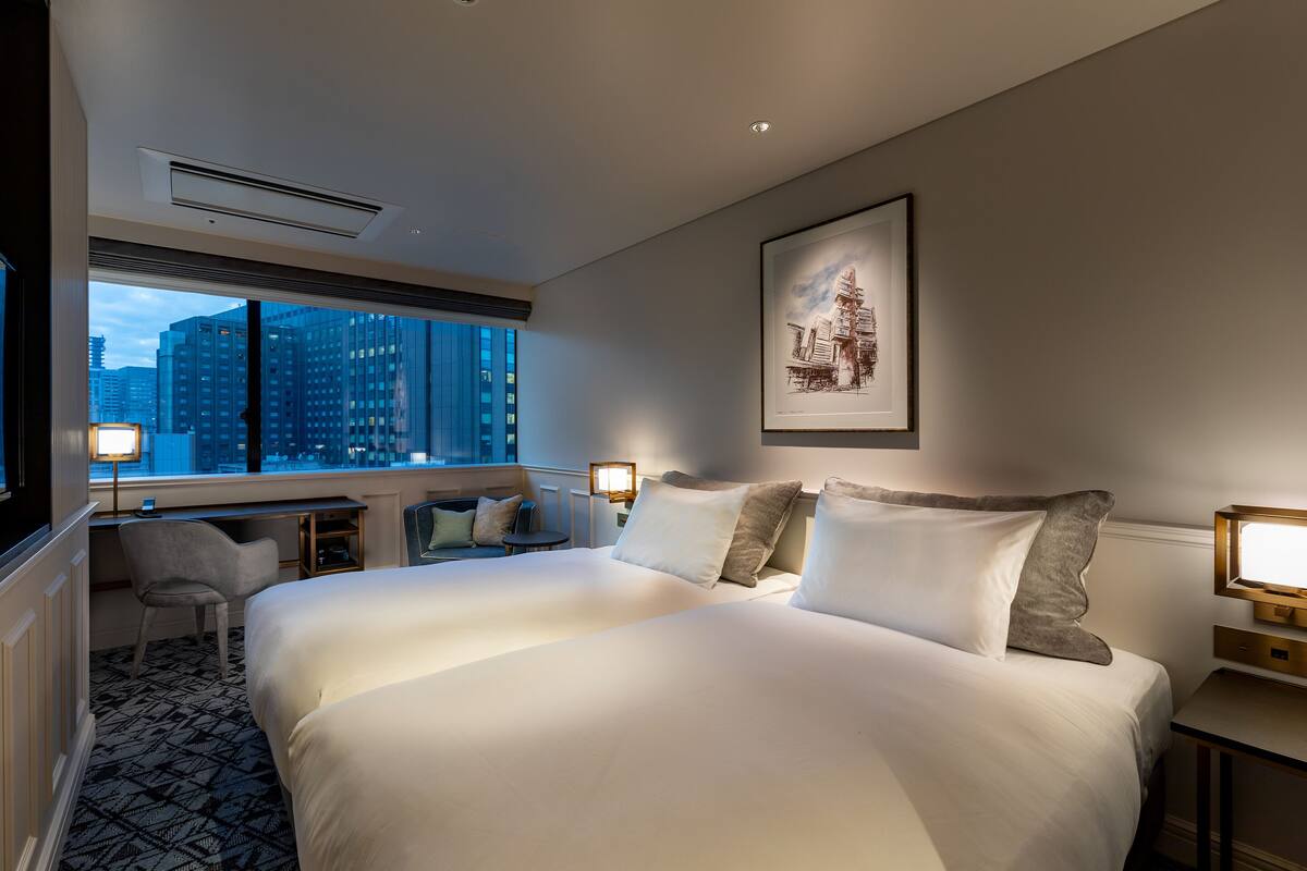 A hotel room with a large bed, a desk, a chair, and a view of tall buildings through the window.