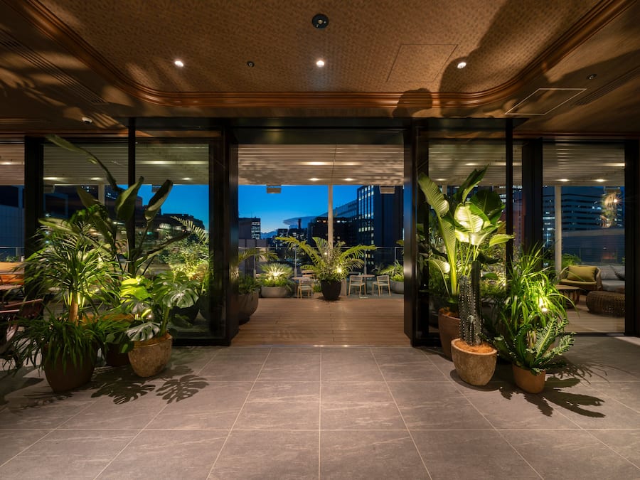 Um lobby de hotel moderno com plantas grandes em vasos, vista para a paisagem urbana e uma área de estar ampla.