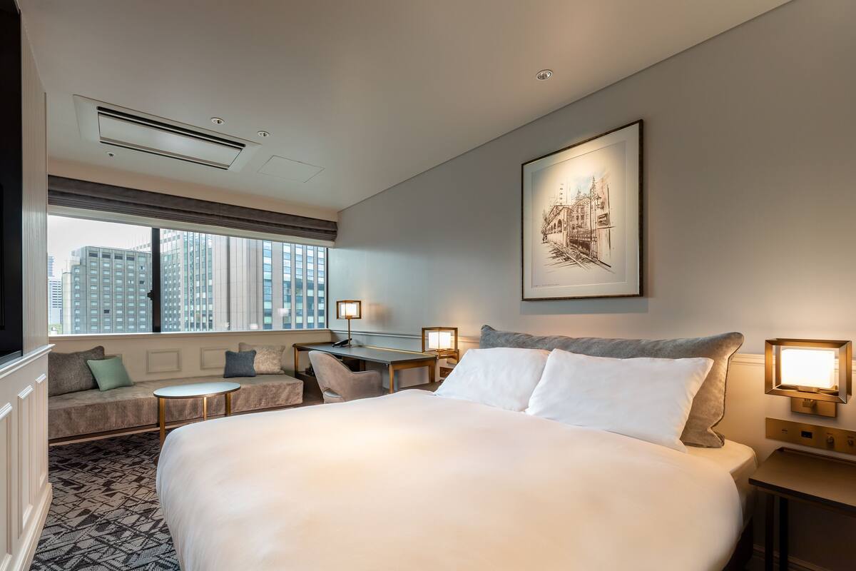 A hotel room with a large bed, a desk, a sofa, and a window with a city view.