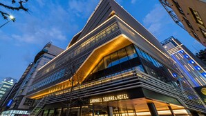 Um hotel moderno com uma placa grande e visível 'GRANBELL SQUARE & HOTEL', com fachada de vidro elegante e uma estrutura de telhado triangular marcante.