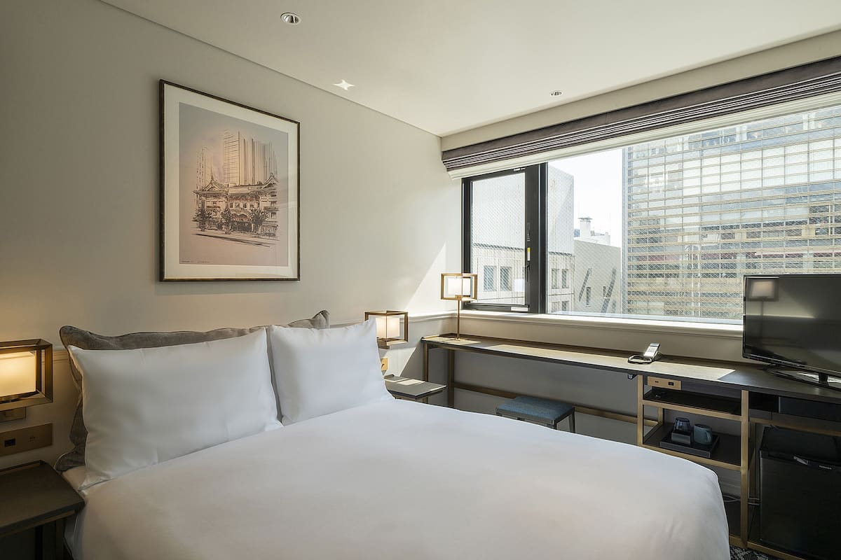 A hotel room with a large bed, a desk with a television, a framed picture on the wall, and a window with a city view.
