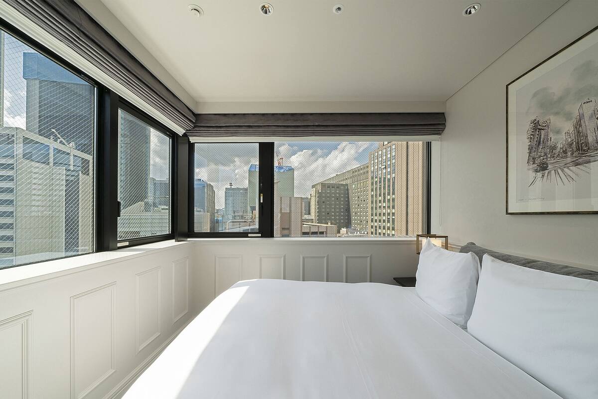 A hotel room with a large bed, a city view through the window, and a framed artwork on the wall.