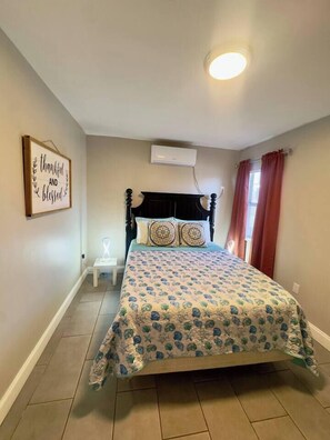 Un dormitorio con cama, aire acondicionado de pared, mesita de noche pequeña y un cartel enmarcados en la pared.