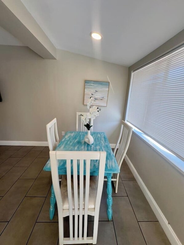 Un coin repas avec une table bleue, des chaises blanches, un tableau au mur et une fenêtre avec des stores.
