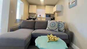 Ein Wohnzimmer mit einem grauen Ecksofa, einem türkisfarbenen Couchtisch und einer Küche im Hintergrund.