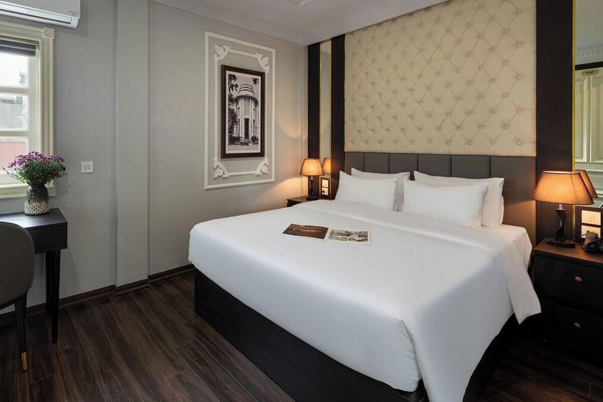 Une chambre d’hôtel avec un grand lit, un bureau avec une chaise, une fenêtre avec des stores, un tableau encadré au mur et une lampe fixée au mur.