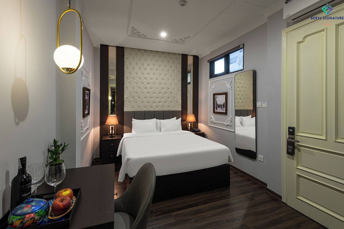 Une chambre d’hôtel avec un grand lit, des lampes de chevet, un bureau avec une bouteille de vin et des verres, et un panier de fruits.