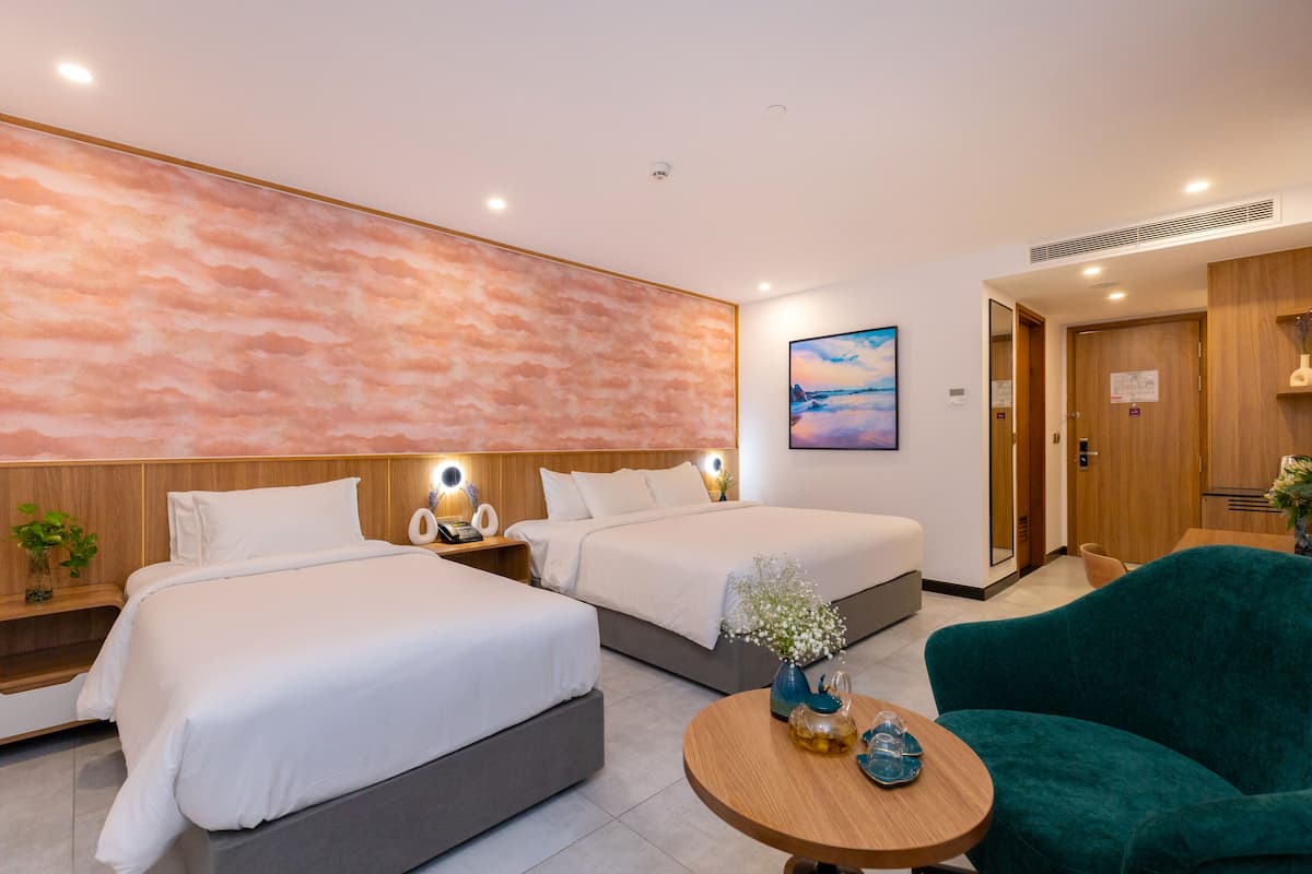 A modern hotel room with two beds, a wooden headboard, a round wooden table, and a teal armchair.