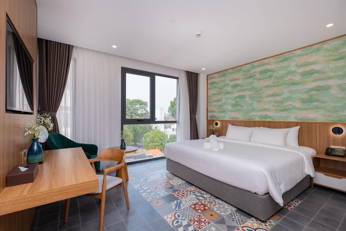 A modern hotel room with a large bed, a desk with a chair, a vase with flowers, and a view of the city through the window.
