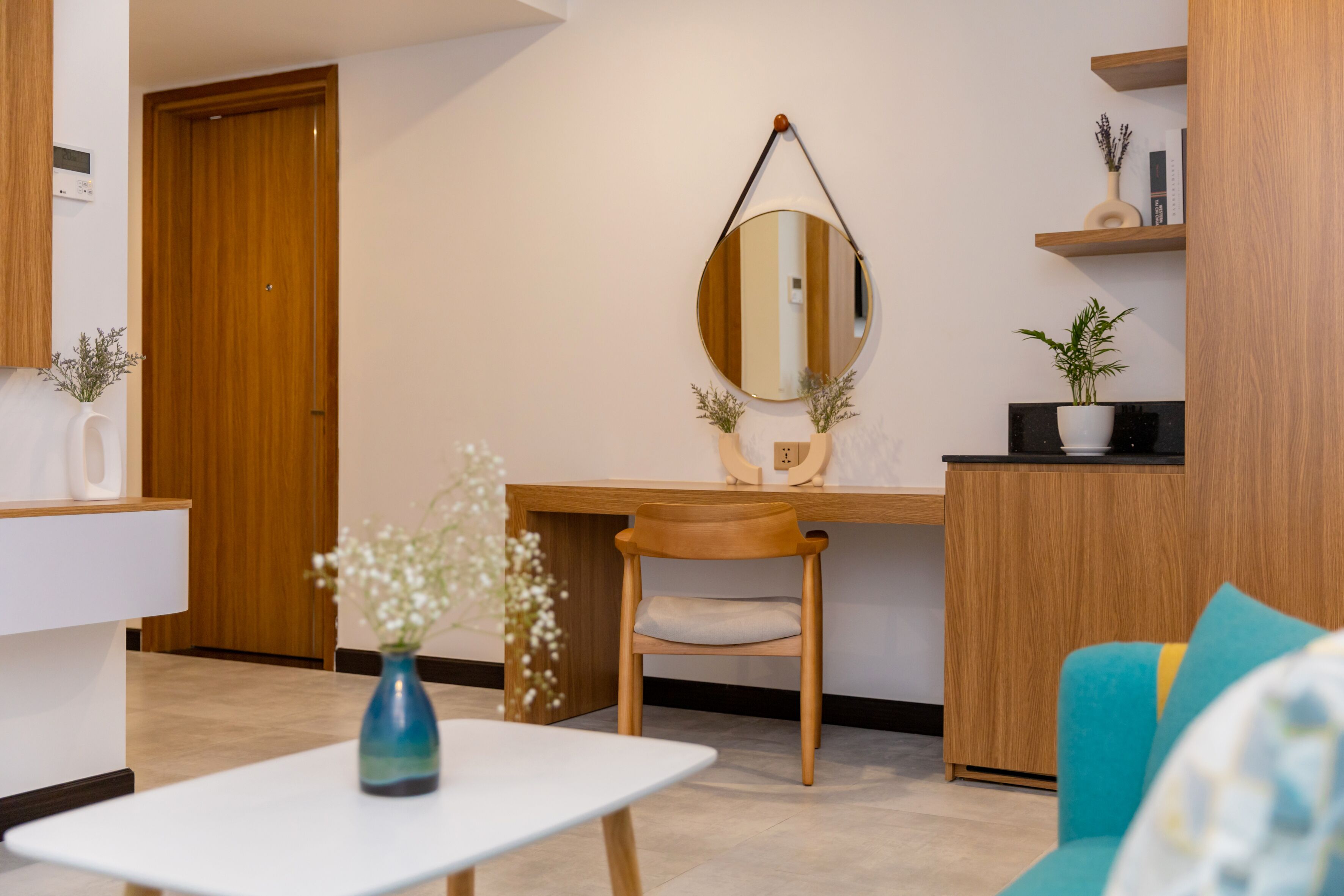 A modern living room with a wooden desk, a round mirror, a blue vase with flowers, and a teal sofa.